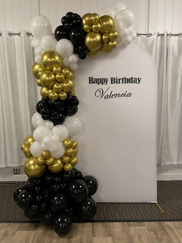 Luxe black, gold, and white organic balloon arch framing a custom "Happy Birthday Valencia" backdrop for a sophisticated Montreal birthday celebration.