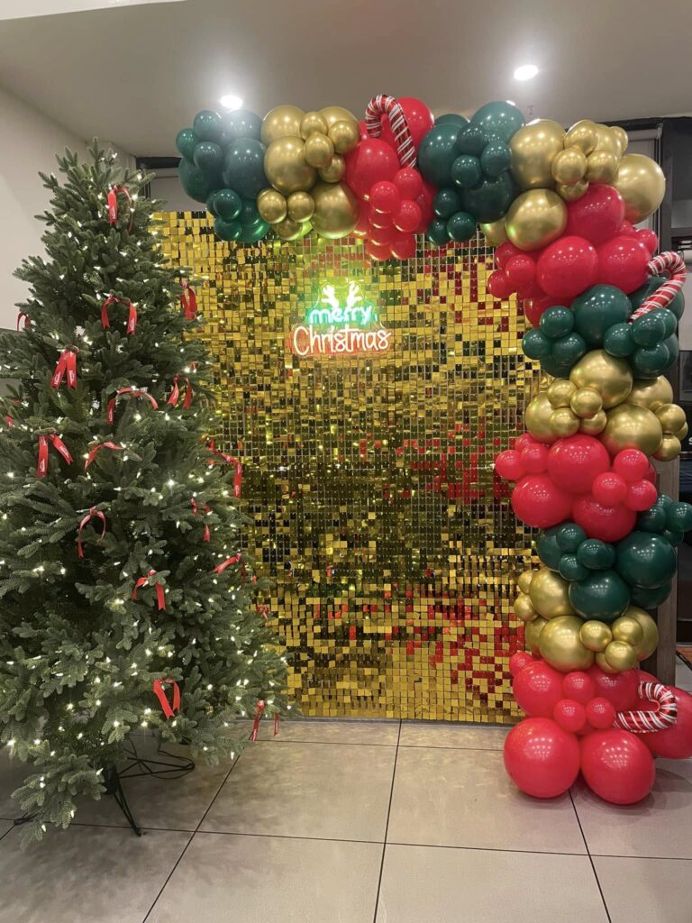Luxe Christmas balloon installation in Montreal featuring a red, gold, and green organic arch with a shimmering gold backdrop and neon "Merry Christmas" signage.