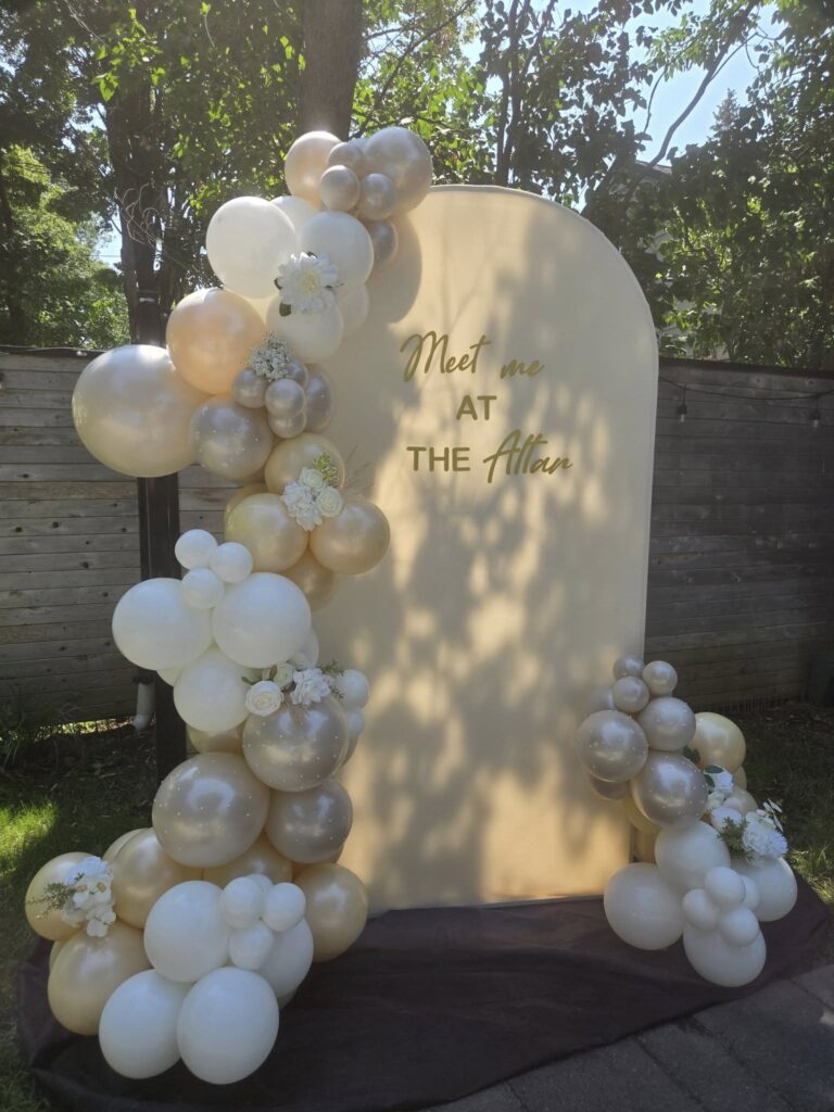 Bespoke wedding balloon garland in champagne and white featuring fresh floral accents and personalized "Meet me at the Altar" backdrop for an outdoor Montreal ceremony.