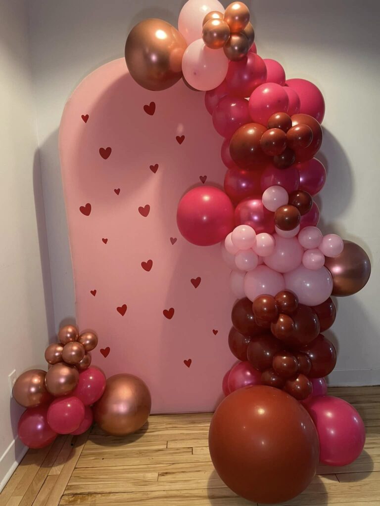 Bespoke organic balloon installation in Montreal featuring clusters of pink, red, and bronze balloons framing a romantic pink heart-patterned arch backdrop.