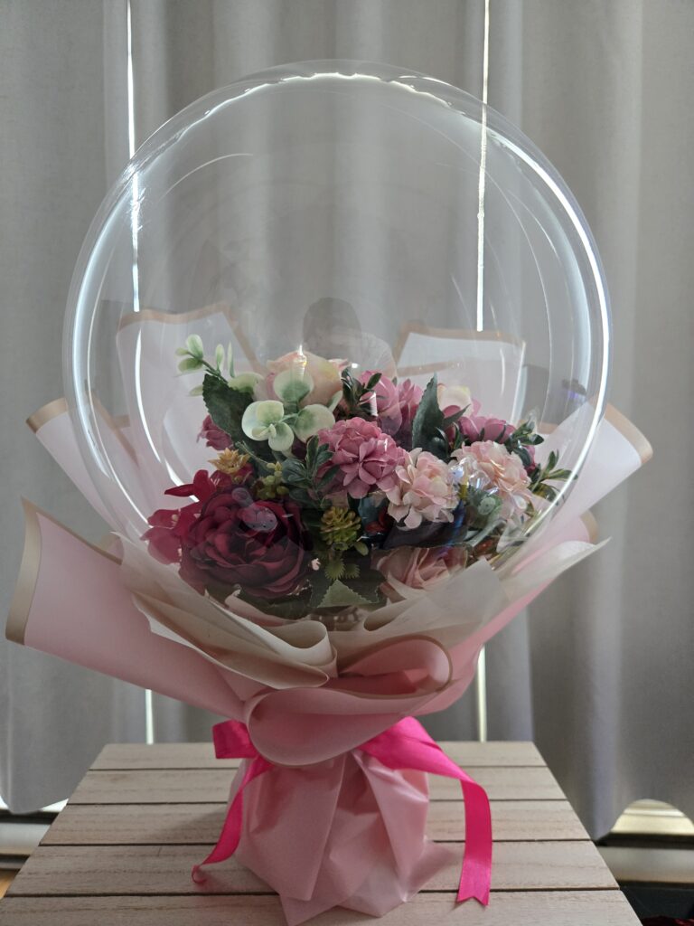 Hand-wrapped Bobo balloon floral bouquet in Montreal featuring vibrant colorful flowers inside a clear balloon, finished with premium pink wrapping and a satin ribbon.