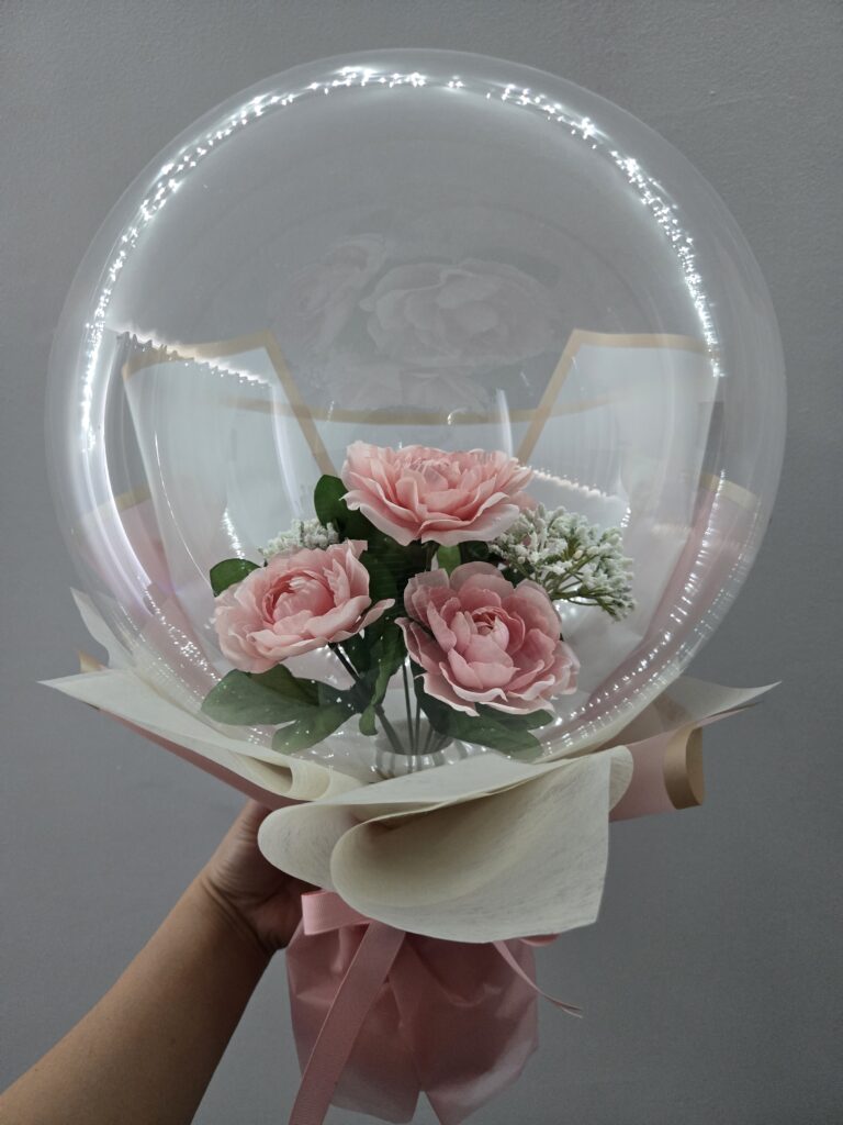 Hand-held artisan Bobo bouquet featuring soft pink roses encapsulated in a clear balloon with elegant white and pink wrapping, a bespoke gift by La Balloonerie Montreal.